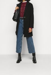 Cappotto in misto lana nero, maglione bordeaux a maglia aperta, jeans blu a vita alta e stivaletti neri con tacco. Borsa nera in mano.