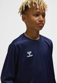 Marinblå långärmad skjorta i slät tyg med rund halsringning och vit "hummel"-logotyp på bröstet. Modellen har kort, ljust hår.