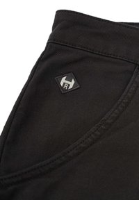 Nahaufnahme von schwarzem Stoff, der eine Tasche mit einem sechseckigen Logo-Patch zeigt, das ein stilisiertes "R" präsentiert. Strukturiert Oberfläche und verstärkte Nähte.