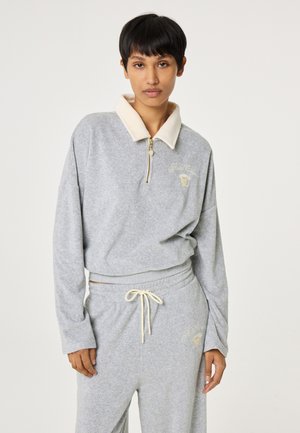 Vrouw met kort donker haar, gekleed in een lichtgrijze fleece trui met crèmekleurige kraag en bijpassende broek met trekkoord, staand tegen een effen achtergrond.