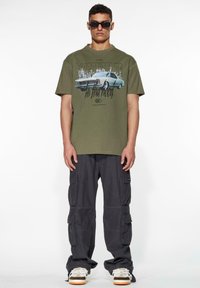 Olivenfarget bomull T-skjorte med et vintage bilmotiv og svart tekst, kombinert med mørke cargo-bukser som har flere lommer og grå joggesko.