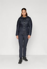 Kvinna står framåt riktad, klädd i svart puffjacka, svarta byxor och svarta vandringsskor mot en enkel ljus bakgrund.