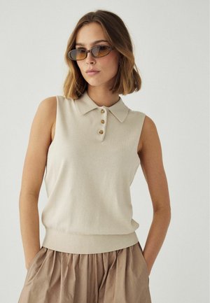 Naine, kes kannab beežikat varrukateta polo särki, helepruuni plisseeritud seelikut ja ovaalseid pruunika varjundiga päikeseprille, seisab käed taskutes ühevärvilise tausta ees.