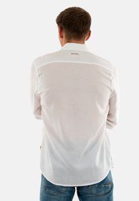 Chemise blanche à manches longues avec une texture lisse, dotée d'un col boutonné et d'un logo à la nuque. Ourlet arrière droit, portée avec un jean.