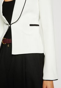 Blazer blanc avec bordure noire et détail de poche, porté sur un top noir, assorti à un pantalon noir et une ceinture bordeaux avec boucle dorée.