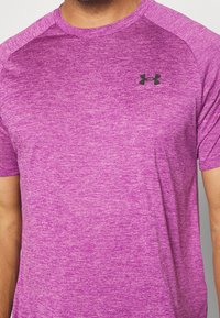 Muž oblečený ve fialovém melírovaném tričku s krátkým rukávem a černým logem Under Armour na levé hrudi.