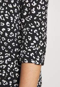 Tissu noir avec un motif de taches de léopard blanches irrégulières sur un revers de manche plié reposant près de l’avant-bras d’une personne.