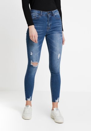 Frau mit blauen, zerrissenen engen Jeans, weißen Sneakern und einem schwarzen langärmeligen Oberteil, steht vor einem einfarbigen hellen Hintergrund.