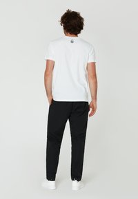 Bílé bavlněné tričko s krátkým rukávem, s kruhovým logem na zádech. Spárováno s černými kalhotami a bílými teniskami. Minimalistický design.