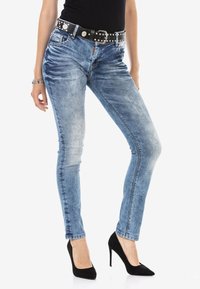 Dunne blauwe jeans met een vervaagde wassing, voorzakken en een zwarte studded riem. Model gecombineerd met zwarte hoge hakken.