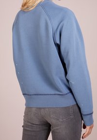 Felpa blu con scollatura e polsini a coste, tessuto testurizzato e lievi macchie di vernice sulle maniche. Abbinata a jeans grigi.