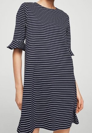 Kobieta ubrana w granatową sukienkę z cienkimi białymi poziomymi paskami oraz krótkimi rękawami z falbankami.