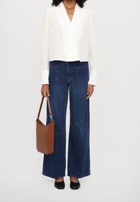 Blusa blanca de manga larga y corta con escote en V. Pantalones vaqueros anchos azul oscuro con bolsillos con parche. Bolso marrón con correa de hombro. Zapatos planos negros.