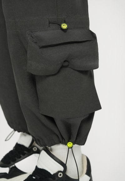 Παντελόνια cargo σε σκούρο γκρι ύφασμα, με μεγάλη πλαϊνή τσέπη, ελαστικές μανσέτες και κίτρινο κορδόνι με λεπτομέρεια χαμογελαστού προσώπου.