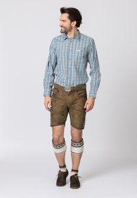 Uomo che indossa una camicia a maniche lunghe a scacchi blu e verde, pantaloni corti in pelle marrone, calze di maglia bianche fino al ginocchio e scarpe scure.