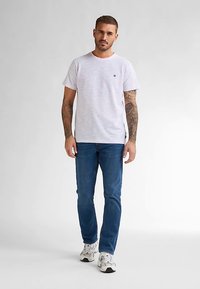 Maglietta a righe bianche con scollo rotondo, indossata con jeans blu e sneakers bianche. Presenta un piccolo logo sul petto.