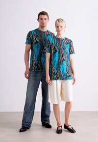 To modeller bærer matchende t-shirts med korteærmer i et farverigt abstrakt mønster med nuancer af blå, rød og gul. Den mandlige model kombinerer det med jeans; den kvindelige model har hvide shorts på.