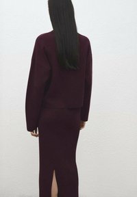 Costum visiniu din două piese, compus dintr-un top scurt cu mâneci largi și o fustă cu o despicătură. Materialul pare moale și texturat.