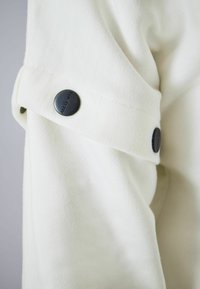 Veste en mélange de coton blanc avec une finition texturée, dotée d'un détail à bouton-pression noir sur la manche. Design simple et moderne.