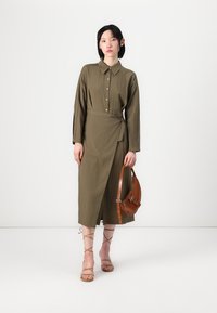 Olive zelené šaty v košovom štýle s dlhými rukávmi, zavinovacím dizajnom a gombíkovými detailmi. Skombinované s hnedou taškou a béžovými sandálmi na remienky.