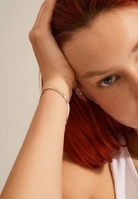 Artimo plano asmuo, kurio veidas ir ranka, puikuojasi plona metaline apyranke, raudonais plaukais ir odos tonais su subtiliais tekstūriniais atspalviais bei nedideliais defektais.