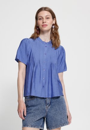 Ung kvinde iført blå kortærmet knapbluse og denimshorts, står mod en ensartet lys baggrund.