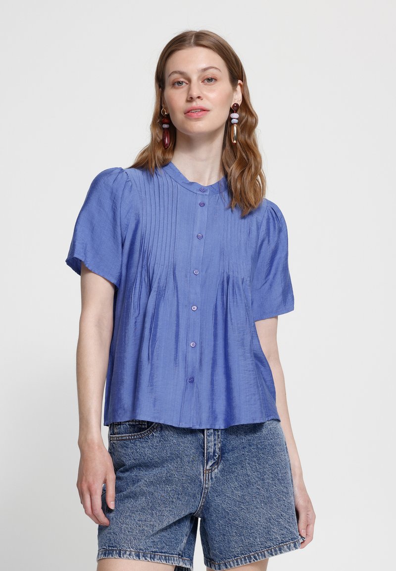 Jeune femme portant un chemisier bleu à manches courtes boutonné et un short en denim, debout devant un fond clair uni.