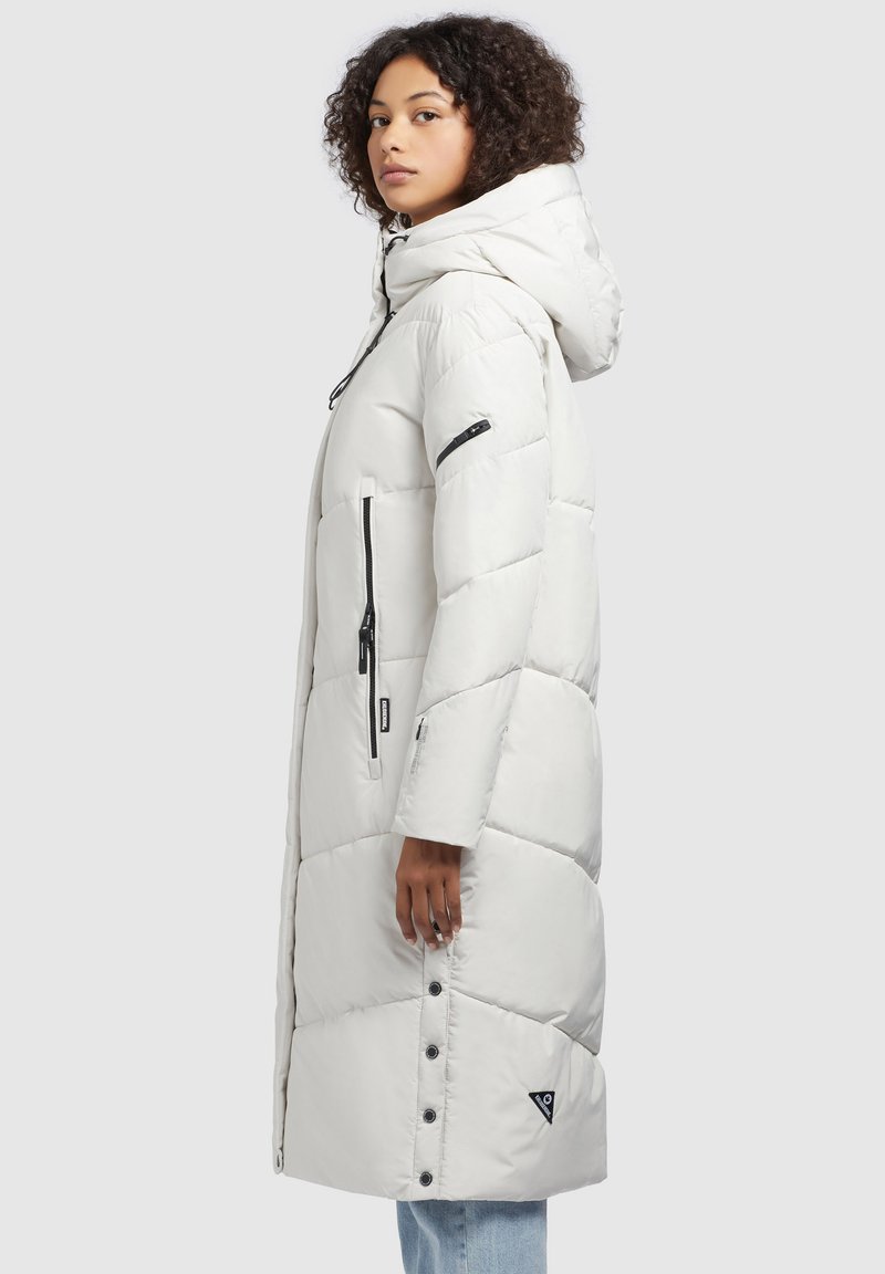 Langer weißer Puffer-Mantel mit Kapuze, ausgestattet mit einem gesteppten Design, Reißverschluss-Seitentaschen und einem schwarzen Logo-Patch am Saum.