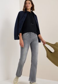 Donna che indossa una giacca blu navy sopra una felpa con cappuccio nera, jeans grigi e sneakers beige, che tiene una borsa riutilizzabile verde oliva in una stanza neutra.