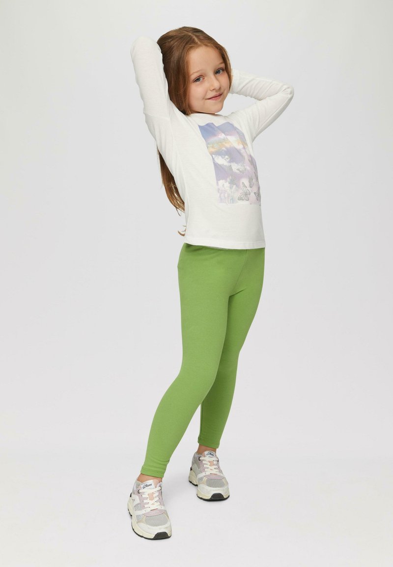 Ein Kind trägt ein weißes langärmliges Shirt mit einem Schmetterlingsmotiv und grüne Leggings, kombiniert mit grauen Turnschuhen. Glatte Stoffe, bequemer Schnitt.