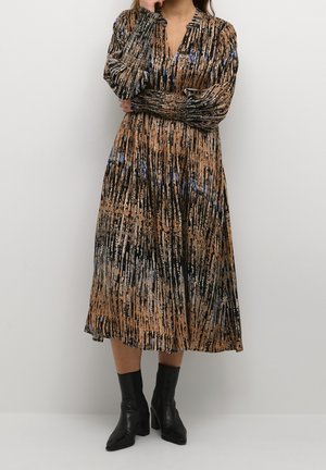 Femme portant une robe midi à manches longues, à motifs, avec un mélange de couleurs marron, noir et bleu, des bottines noires à talons.