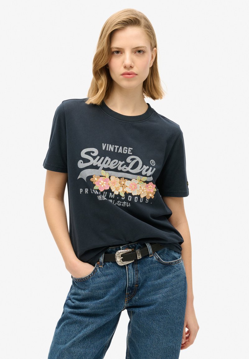 T-shirt in cotone blu navy con un design floreale ricamato in rosa e giallo, stampa "SuperDry" in argento e colletto girocollo classico.