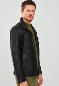 Giubbotto di pelle nera con un design strutturato, dotato di colletto alto, chiusura frontale con zip e due tasche laterali. Texture liscia e vestibilità su misura.