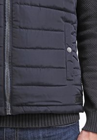 Granatowa pikowana kurtka z przednim zamkiem błyskawicznym, z boczną kieszenią na zatrzask. Rękawy i dół wykończone ściągaczowym, szarym dzianym materiałem.