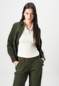Veste en velours côtelé vert olive sur un polo crème à côtes, associée à un pantalon en velours côtelé vert olive assorti avec des boutons et des poches noires.