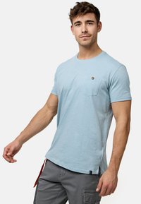 Helles blaues T-Shirt mit kurzen Ärmeln aus weichem Stoff, ausgestattet mit einer Brusttasche und einem kleinen Logo-Detail in der Nähe des Ausschnitts.