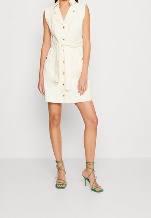 Ärmelloses cremefarbenes Kleid mit Knöpfen und Gürtel, knielang, getragen mit grünen, mit Riemchen versehenen Sandaletten mit Absatz an einer stehenden Person.