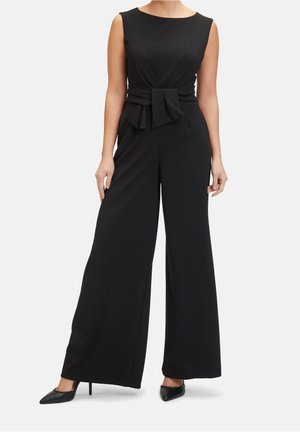 Combinaison noire sans manches avec taille cintrée et détail nœud, dotée de pantalons à jambes larges et d'une texture de tissu lisse. Associée à des talons noirs.