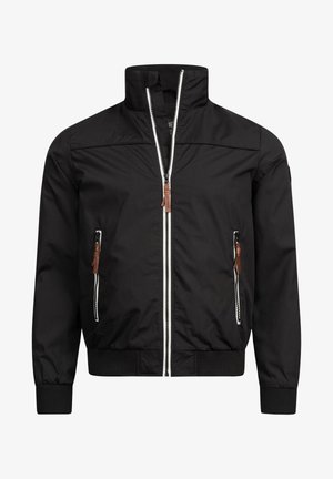 Zwarte bomberjack met rits aan de voorkant, elastische manchetten en een bruine leren patch op de mouw. Gecombineerd met donkerblauwe jeans en witte sneakers.