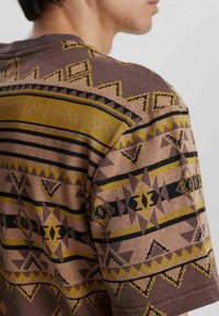 Camisa de punto de manga corta en marrón con patrones geométricos en negro y amarillo. Presenta rayas diagonales y una textura en la tela.