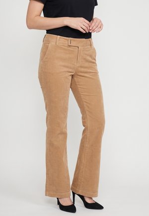 Donna che indossa pantaloni di velluto a coste beige, top nero e scarpe nere con tacco alto, in piedi con le mani giunte vicino alla vita.