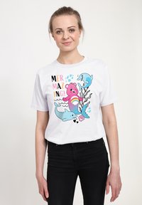 Camiseta blanca con un gráfico colorido de un oso rosa, delfines y el texto que dice "¡MERMAZING!" sobre un fondo simple.