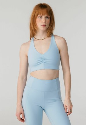 Žena s červenými vlasmi, oblečená v svetlomodrom športovom podprsenke a legínach s vysokým pásom, stojaca pred jednoduchým sivým pozadím.