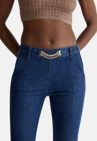 Pantaloni in denim blu scuro con un design aderente, caratterizzati da un accent in catena metallica sulla vita e tasche laterali. Maglia a maglia testurizzata sopra.