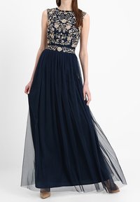 Kvinna i en ärmlös marinblå klänning med beige blommig broderi på bysten och en flytande genomskinlig kjol, håller i klänningens sidor.