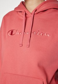 Hoodie rosa con tasca frontale, con logo "Champion" ricamato. Presenta un cappuccio con cordini e un tessuto morbido e texture.