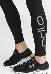 Leggings sportivi neri con un dettaglio del logo argentato sul lato, abbinati a scarpe da corsa nere in mesh con suola bianca e accenti grigi.