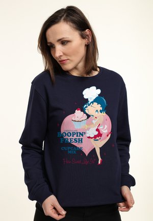 Marineblå sweatshirt med en grafik af en tegneseriefigur i en kokkehue, der holder en cupcake, med farverige tekst og accenter.