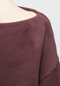 Artimas kadras, kuriame matyti žmogus vilkintis laisvą, tamsiai raudoną, minkštos medžiagos viršų su plačiu iškirpimu, atidengiančiu viršutinę nugaros ir peties dalį.