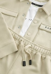 Beige corduroy takki, jossa on nepparit ja päällyspocketti, yhdistettynä samaan sävyyn piilotettuihin housuihin, joissa on logotunnisteet ja sileä pinta.